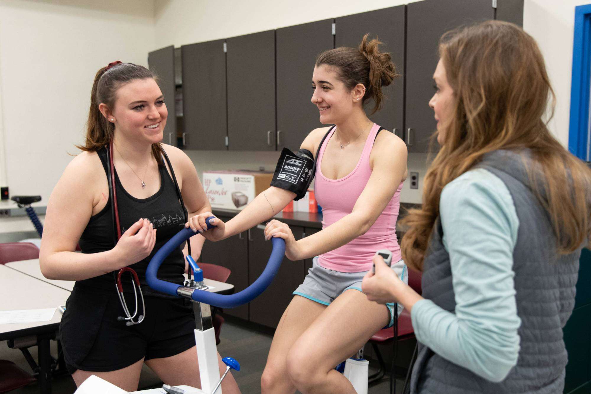 Women in the Health, Wellness, and Movement House happily work together on a project in a fitness lab. One rides a stationary bike with a blood pressure cuff, while the others, smiling, observe and chat.